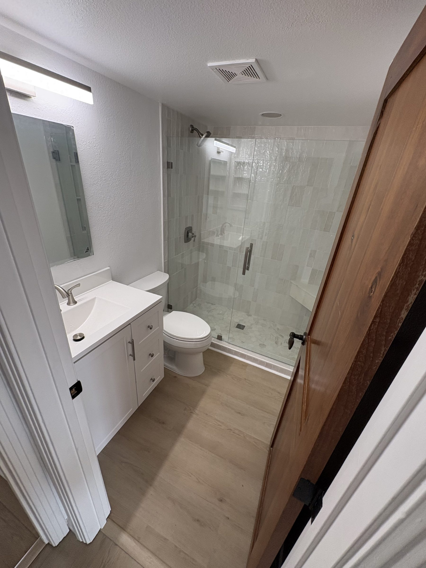 Finished bathroom with a white vanity, wide mirror with a light bar above it, toilet and a tiled walk-in shower with a glass door, over plank flooring.